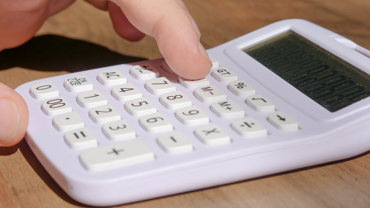 hero-img A hand pressing keys on a white calculator placed on a wooden desk in bright daylight.